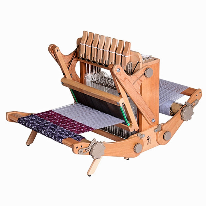 The Ashford Katie Table Loom, crafted from natural wood, features a weaving width of 12 inches. Pictured in use, the loom displays its heddle frame raised, with black and red woven fabric emerging, showcasing the loom's functionality and compact design, perfect for both beginners and experienced weavers looking for a portable weaving solution.